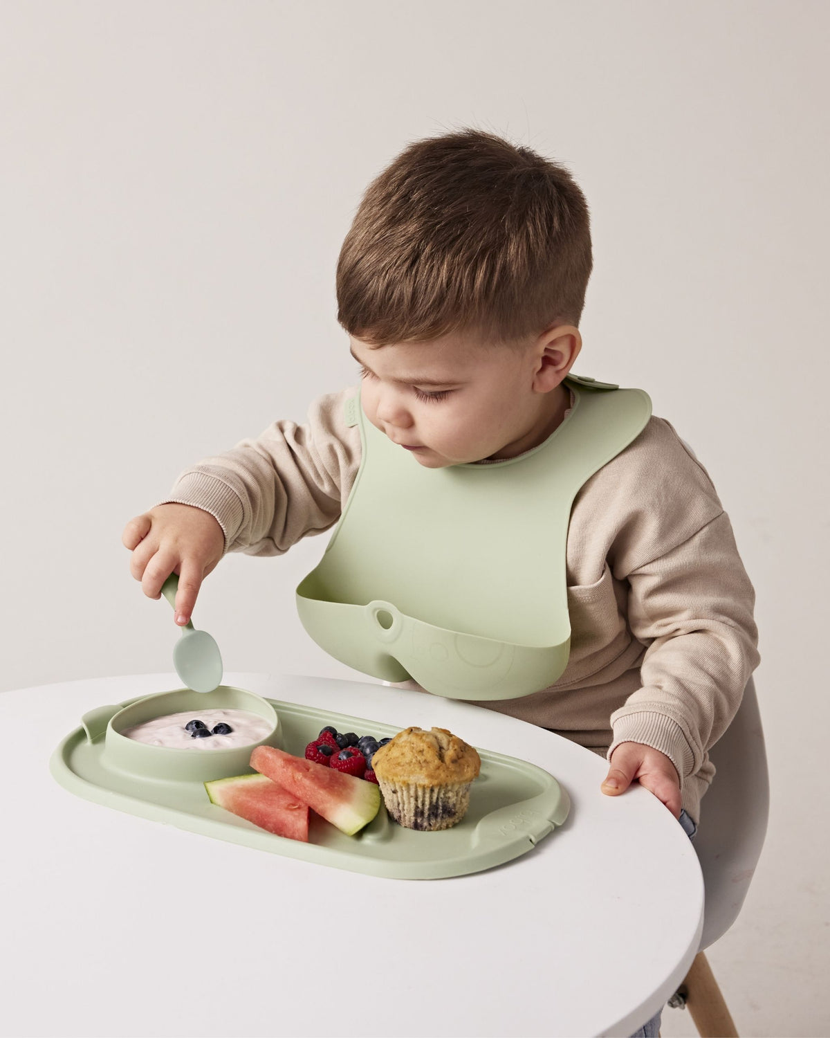 同色のトレーとスプーンで食事をする、シリコンビブを着けた男の子